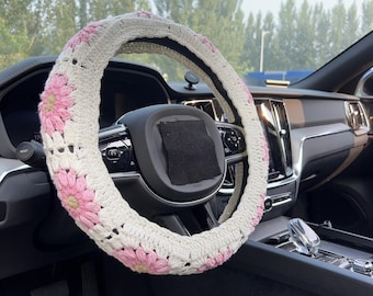 May include: A white crochet steering wheel cover with pink daisy flower accents. The cover is installed on a black steering wheel inside a car. The interior of the car is visible in the background.