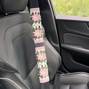 May include: Crocheted seat belt covers in a car. The covers feature a floral design with pink, white, and green colors. The covers are attached to a black seat belt, and the car seat is black.