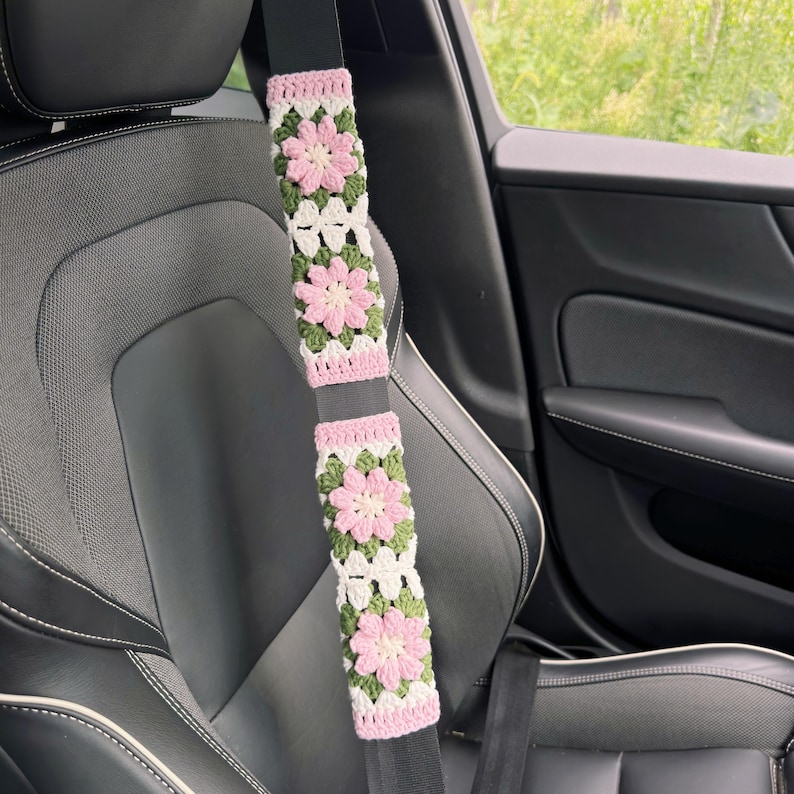 May include: Crocheted seat belt covers in a car. The covers feature a floral design with pink, white, and green colors. The covers are attached to a black seat belt, and the car seat is black.