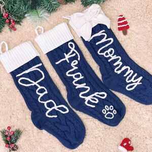 May include: Three navy blue Christmas stockings with white accents. The stockings are cable-knit and feature the names "Dad", "Frank", and "Mommy" in white script. One stocking has a paw print. A white bow adorns one stocking.