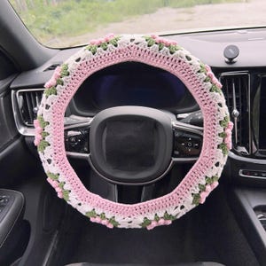 May include: A crocheted steering wheel cover in pink, white, and green. The cover is made of yarn and features a floral design. The cover is shown on a black steering wheel in a car.