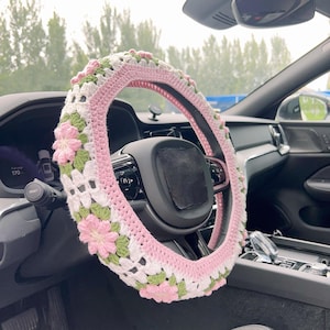 May include: A crocheted steering wheel cover in a car interior. The cover features a pink, white, and green floral design. The steering wheel is black, and the car's dashboard is visible in the background.