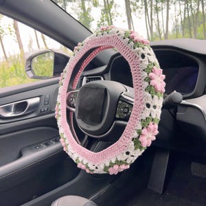 May include: A pink, white, and green crochet steering wheel cover with a floral design. The cover is installed on a black steering wheel inside a car. The crochet pattern includes pink flowers and green leaves.
