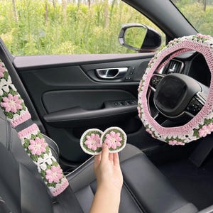 May include: Crocheted car accessories set in pink, white, and green floral pattern. Includes a steering wheel cover, seat belt covers, and two coasters. The accessories are designed to fit a car interior.