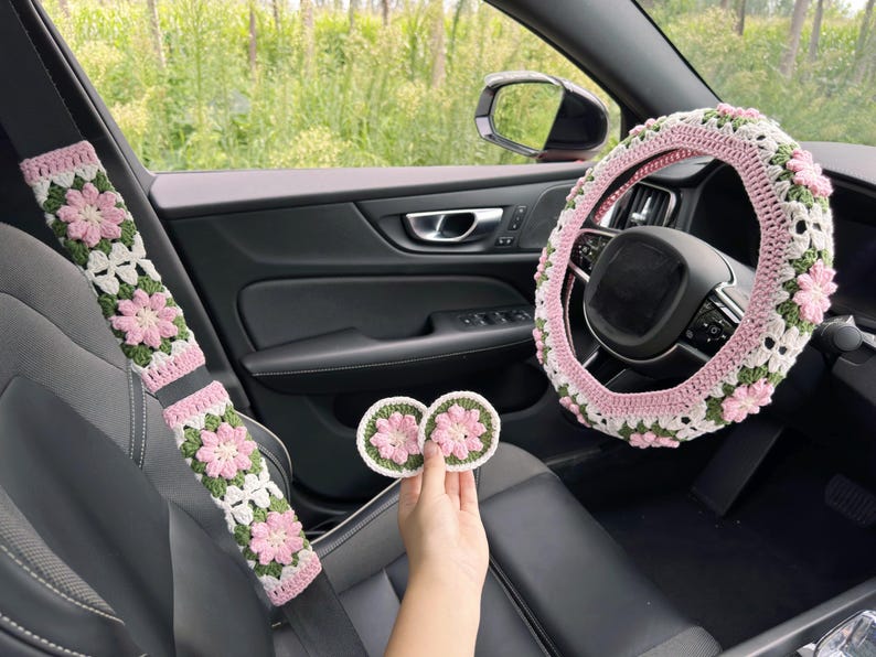 May include: Crocheted car accessories set in pink, white, and green floral pattern. Includes a steering wheel cover, seat belt covers, and two coasters. The accessories are designed to fit a car interior.