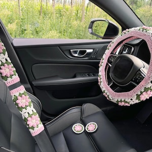 May include: Crocheted car accessories set in a car interior. The set includes a steering wheel cover, seat belt covers, and two round coasters. The items feature a pink, white, and green floral pattern, adding a decorative touch to the car.
