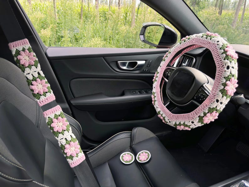 May include: Crocheted car accessories set in a car interior. The set includes a steering wheel cover, seat belt covers, and two round coasters. The items feature a pink, white, and green floral pattern, adding a decorative touch to the car.