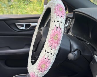 May include: A white crochet steering wheel cover with pink daisy flower accents. The cover is installed on a black steering wheel inside a car. The crochet design adds a decorative touch to the vehicle's interior.