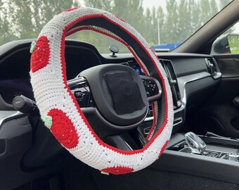 May include: A white and red crocheted steering wheel cover with a strawberry design. The cover is installed on a black steering wheel inside a car. The strawberry design features red fruit with green leaves.