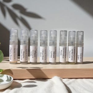 May include: Eight miniature perfume sample bottles are displayed. Each clear bottle features a white label with the brand name "NINETEEN" and a fragrance name, such as "Cherry Rose" or "Garden of Eden". The bottles are arranged on a light wooden surface.