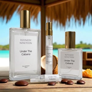 May include: Several clear glass perfume bottles with gold caps are arranged on a wooden surface. The bottles are labelled "REFINERY NINETEEN" and "Under The Cabana." A coconut and small, brown, oval-shaped objects are also visible.