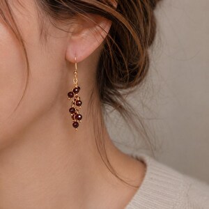 Peut inclure: Boucles d'oreilles pendantes dorées avec des grappes de petites perles rouge foncé. Les boucles d'oreilles sont suspendues à de délicats crochets dorés, créant un effet en cascade. La personne porte un pull crème et a les cheveux bruns.