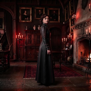 May include: A floor-length black leather coat featuring a high collar, long sleeves with studded detailing along the shoulders and cuffs, and vertical red piping accents running down the front and back panels of the garment.
