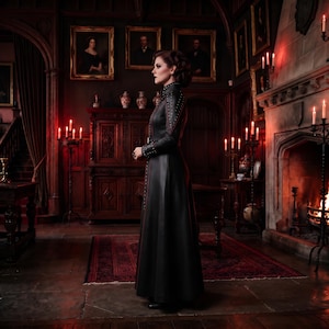 May include: A floor-length black leather coat featuring a high collar, long sleeves, and vertical rows of silver studs along the front placket and shoulders. The coat has a fitted bodice that transitions into a full-length skirt with a smooth finish.