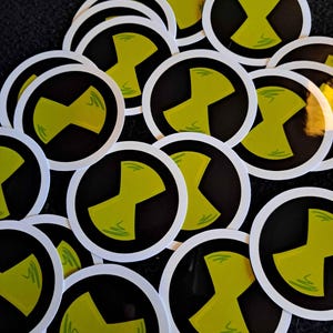 May include: A collection of round stickers featuring a black circle with a lime green and white design. The stickers have a white border and are scattered across a dark surface. The design is a stylized X shape.