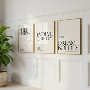 May include: Three framed art prints with gold frames and white backgrounds. The prints feature black text with motivational phrases: "BUILT DIFFERENT.", "EVOLVE QUIETLY.", and "DREAM BOLDLY.". The prints are displayed on a white wall.