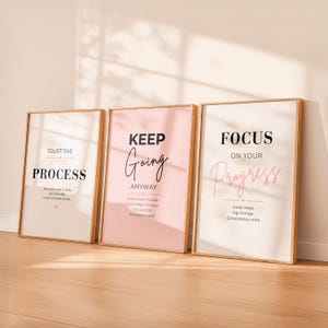 Puede incluir: Tres impresiones artísticas enmarcadas con marcos dorados. Las impresiones presentan citas inspiradoras en texto negro y rosa sobre fondos blancos y rosas. Las citas incluyen "Trust the Process", "Keep Going Anyway" y "Focus on Your Progress".