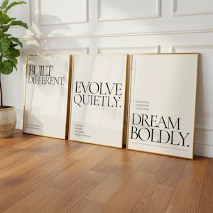 May include: Three framed art prints with gold borders lean against a wall. The prints feature black text on a cream background. The text reads "BUILT DIFFERENT", "EVOLVE QUIETLY", and "DREAM BOLDLY".