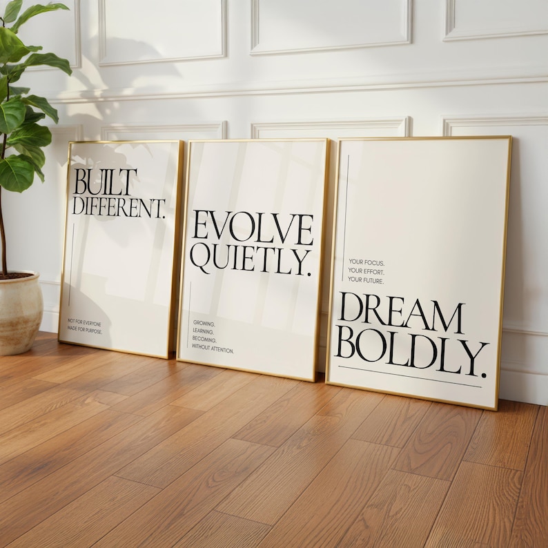 May include: Three framed art prints with gold borders lean against a wall. The prints feature black text on a cream background. The text reads "BUILT DIFFERENT", "EVOLVE QUIETLY", and "DREAM BOLDLY".