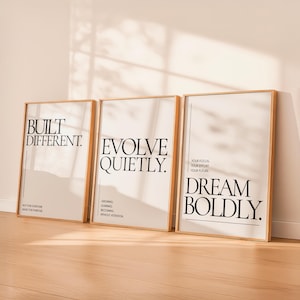 May include: Three framed art prints with motivational quotes. The prints have a light beige background and black text. The quotes read "BUILT DIFFERENT", "EVOLVE QUIETLY", and "DREAM BOLDLY". The frames are a light wood color.
