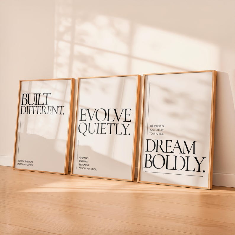 May include: Three framed art prints with motivational quotes. The prints have a light beige background and black text. The quotes read "BUILT DIFFERENT", "EVOLVE QUIETLY", and "DREAM BOLDLY". The frames are a light wood color.