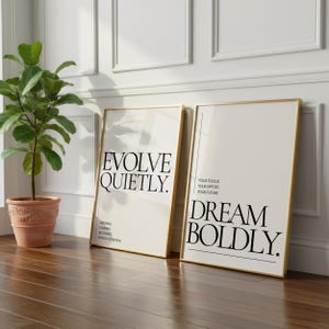 May include: Two framed art prints with gold trim lean against a white wall. One print reads "EVOLVE QUIETLY" and the other says "DREAM BOLDLY." A potted plant sits to the left on a wooden floor.