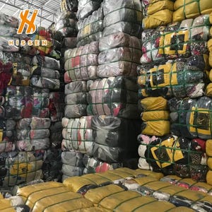 Puede incluir: Un gran almacén lleno de numerosos fardos rectangulares de ropa variada, cada uno fuertemente comprimido y envuelto en film plástico transparente o amarillo, asegurado con flejes de plástico verde y apilado en columnas altas.