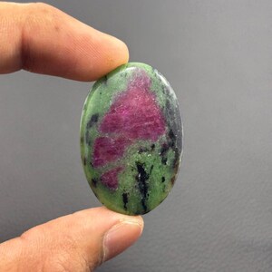 May include: An oval-shaped gemstone with a vibrant mix of colors. The stone features a striking combination of deep red, green, and black hues. The polished surface of the stone reflects light, highlighting the natural patterns and textures. This gemstone is a unique piece for jewelry or collection.