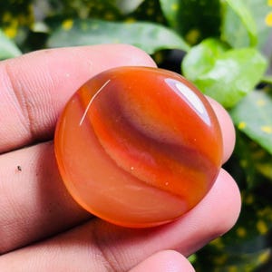 Könnte beinhalten: Ein polierter, runder Edelstein mit einem lebendigen orangefarbenen und rotbraunen Bandmuster. Der Stein wird in einer Hand gehalten, mit grünem Laub im Hintergrund. Der Edelstein hat eine glatte, glänzende Oberfläche.