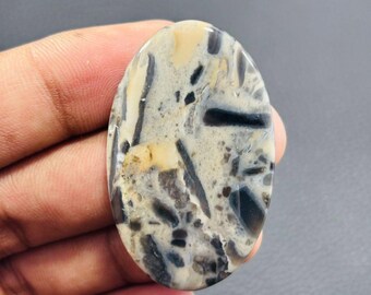 May include: Oval-shaped gemstone with a unique pattern. The stone features a mix of colors, including beige, gray, and black, with a marbled appearance. The polished surface showcases a natural, organic design, suitable for jewelry or crafting.