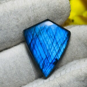 May include: A vibrant blue gemstone with a diamond shape. The stone has a polished surface with internal lines and patterns. The gemstone is held in a white fabric. The background is a blurred yellow.