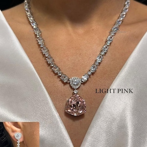 Pode incluir: Um colar de tom prateado com uma série de pedras claras de corte oval que levam a um pingente triangular central com uma grande pedra rosa claro. Um brinco a condizer com uma pedra clara e uma gota triangular rosa claro também é mostrado. Texto: LIGHT PINK e N-1540.