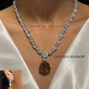 Pode incluir: Um conjunto de colar e brincos com corrente prateada e pedras transparentes de corte redondo. O colar tem um pingente triangular central com uma grande pedra facetada castanho-dourada. Texto: HK PREMIUM QUALITY, GOLDEN SHADOW, N-1540.
