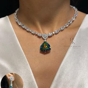 Pode incluir: Um colar de tom prateado com uma série de pedras claras de corte oval e um pendente central triangular com uma gema iridescente multicolorida. Um brinco a condizer com uma pedra clara e um pendente iridescente é mostrado no canto.