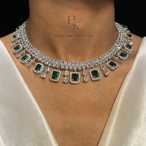 Peut inclure: Un collier argenté orné de pierres vertes émeraude et de pierres claires rectangulaires et en forme de goutte. Le collier est autour du cou d'une personne. Le texte "PREMIUM QUALITY" est visible.