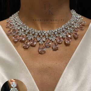 Peut inclure: Un collier imposant composé de pierres transparentes taille brillant disposées en un col dense et multicouche, agrémenté de pierres en forme de poire couleur pêche. Le bijou porte le texte HK PREMIUM QUALITY Feel Like A Diamond.