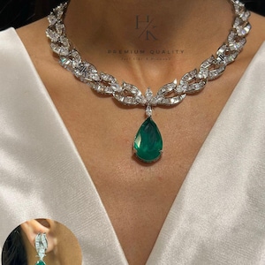 Op de afbeelding: Een zilveren ketting met een druppelvormige smaragd hanger. De ketting heeft een gedetailleerd ontwerp met kleine, heldere stenen. Bijpassende smaragd oorbellen zijn ook zichtbaar. De tekst "PREMIUM QUALITY" is zichtbaar.