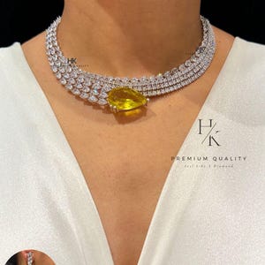 Op de afbeelding: Een sprankelende diamanten ketting met een grote gele peervormige edelsteen hanger. De ketting is meerlaags en voorzien van kleinere, heldere edelstenen. Een bijpassende oorbel is ook zichtbaar. De tekst "HK PREMIUM QUALITY".