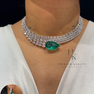 Op de afbeelding: Een diamanten ketting met een grote, peervormige smaragdgroene hanger. De ketting is gemaakt van meerdere rijen kleine diamanten. Een bijpassende oorbel is links onderin afgebeeld. De tekst "HK PREMIUM QUALITY feel like a Diamond" is zichtbaar.
