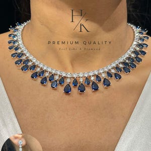 Puede incluir: Un collar plateado y un juego de aretes a juego con piedras azules en forma de lágrima y piedras de acento transparentes. El collar tiene una fila de piedras transparentes y una fila de gotas azules. El texto "PREMIUM QUALITY Feel Like A Diamond" es visible.