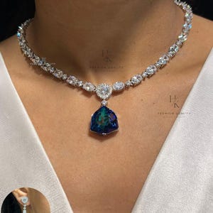 Pode incluir: Um colar de tom prateado com uma série de pedras transparentes de corte redondo ao longo da corrente e um grande pendente triangular azul iridescente no centro. Um brinco a condizer com uma pedra azul semelhante é mostrado no canto inferior.