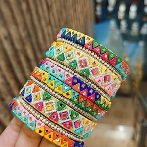 May include: A collection of vibrant bangles with detailed geometric designs. The bangles display a range of bright colours, such as yellow, pink, blue, and green, with gold and silver detailing. The design incorporates small, sparkling embellishments.