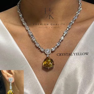 Pode incluir: Um colar de tom prateado com uma fileira de pedras transparentes de corte redondo e um grande pingente de cristal amarelo facetado. O colar inclui uma cravação em forma de coração acima do pingente. Um brinco a condizer com cristal amarelo é exibido.