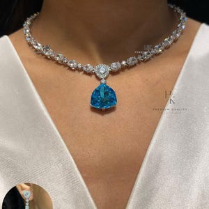 Pode incluir: Um colar de tom prateado com um pingente central de pedra preciosa azul brilhante triangular rodeado por pequenas pedras claras, com uma corrente de pedras claras alternadas. Um pequeno círculo mostra um brinco a condizer com o texto N-1540.