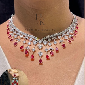 Op de afbeelding: Een diamanten ketting en bijpassende oorbellen set. De ketting heeft een reeks heldere, rond geslepen diamanten en druppelvormige rode edelstenen. De oorbellen hebben een vergelijkbaar ontwerp. De tekst "PREMIUM QUALITY" is zichtbaar.