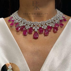 Peut inclure: Un collier argenté orné de nombreuses pierres transparentes à facettes et de plusieurs pierres rose foncé en forme de goutte suspendues au bord inférieur. Une boucle d'oreille assortie est visible dans un encart circulaire. Texte N-1545.