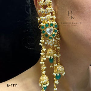 May include: Gold-toned statement earring with emerald green and clear gemstones, and dangling pearl chains. The earring features a floral design and a bell-shaped charm. The text "E-1111" is visible.