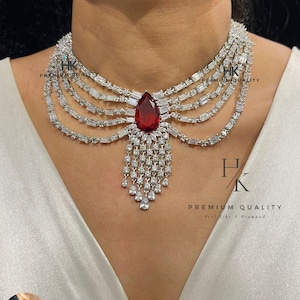 May include: A silver necklace with a large, teardrop-shaped red gemstone pendant. The necklace has multiple layers of baguette-cut stones and smaller, dangling teardrop-shaped stones. The image also includes a small earring with a similar design.