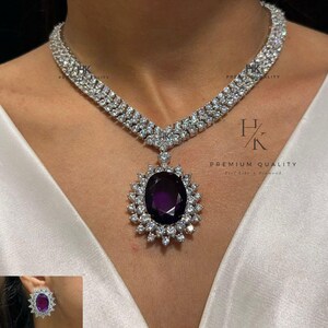 Op de afbeelding: Een schitterende ketting met een grote, ovale, dieppaarse edelsteen hanger, omringd door sprankelende witte stenen. De set bevat bijpassende oorbellen en een ring, allemaal met hetzelfde ontwerp. De tekst "HK PREMIUM QUALITY" is zichtbaar.