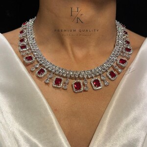 Op de afbeelding: Een prachtige ketting met levendige rode rechthoekige edelstenen, aangevuld met kleinere, heldere stenen. De ketting is gezet in een zilverkleurig metaal, ontworpen om om de hals te zitten. De tekst "PREMIUM QUALITY" is zichtbaar. Een luxe sieraad.